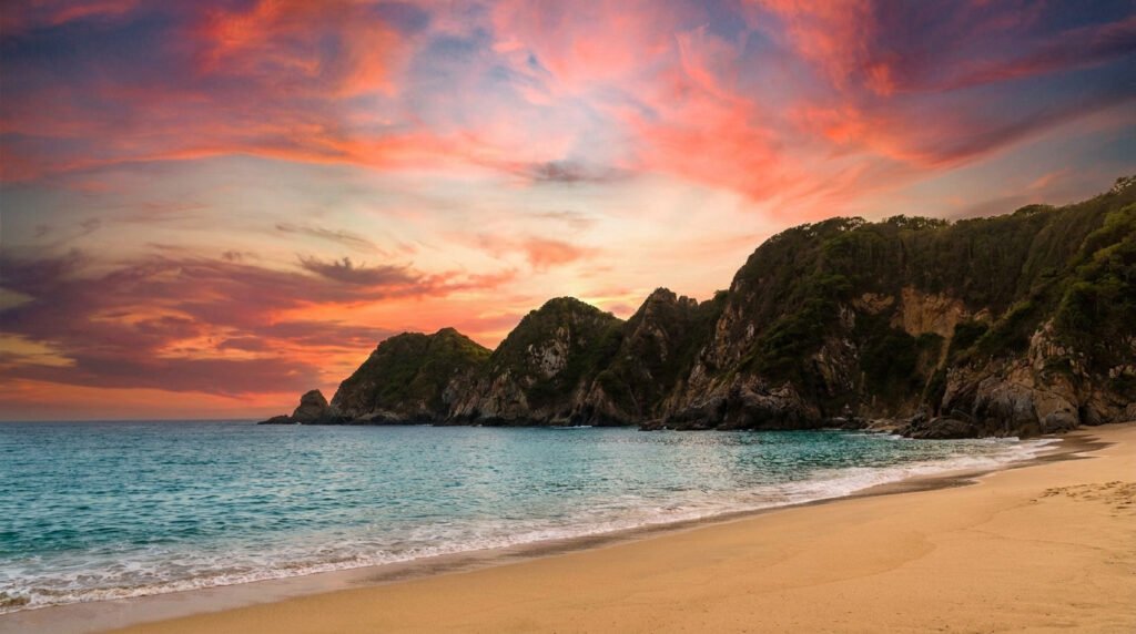 A golden sand beach meets turquoise ocean waters under a vibrant sunset sky with rugged cliffs in the background.